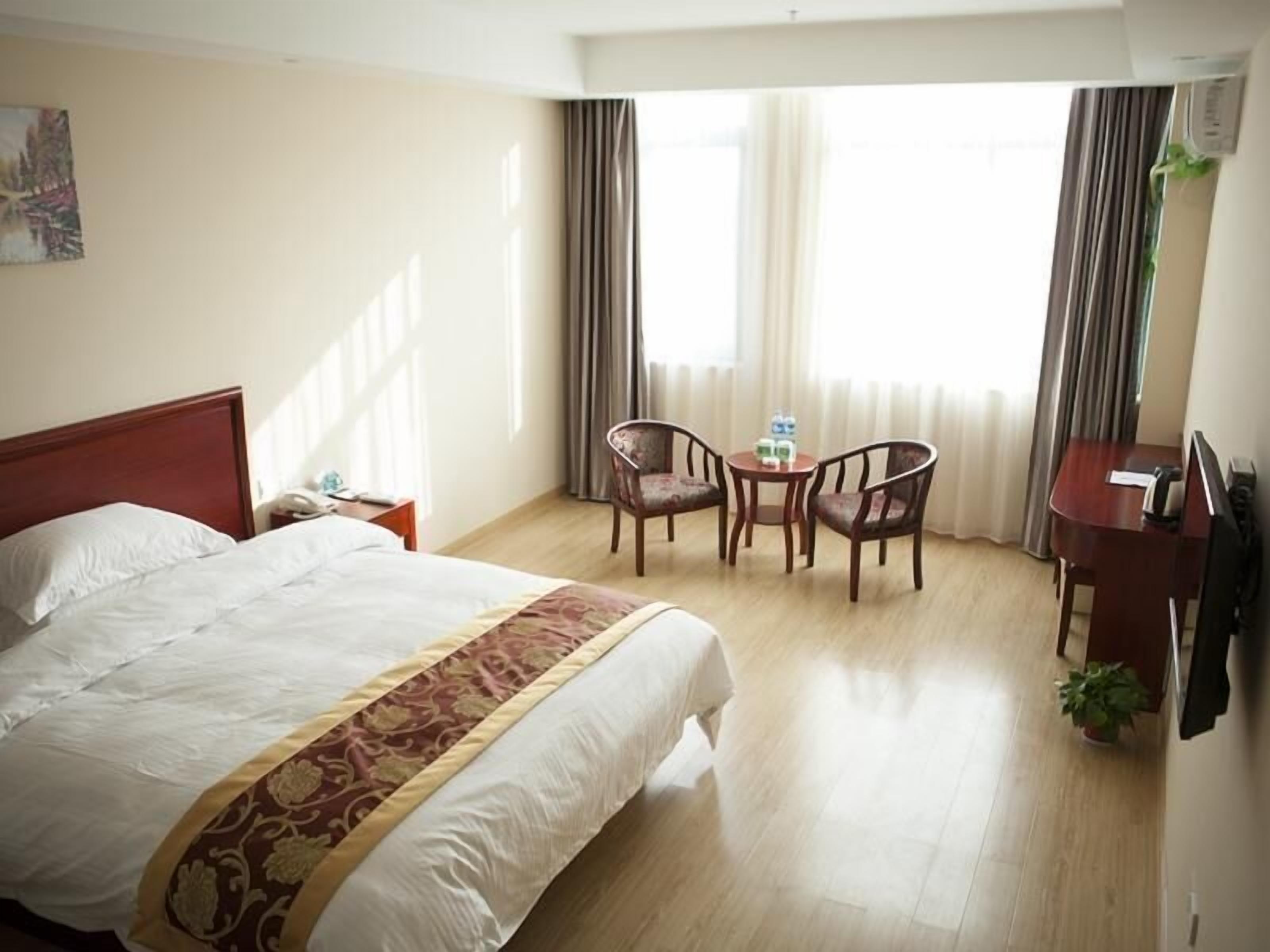 A hotel room with a large bed, a desk, two chairs, a small table with water bottles, and a potted plant.