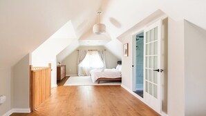 Une chambre spacieuse située dans les combles, avec un plancher en bois, un lit, une fenêtre avec des rideaux et une porte dotée de panneaux en verre dépoli.