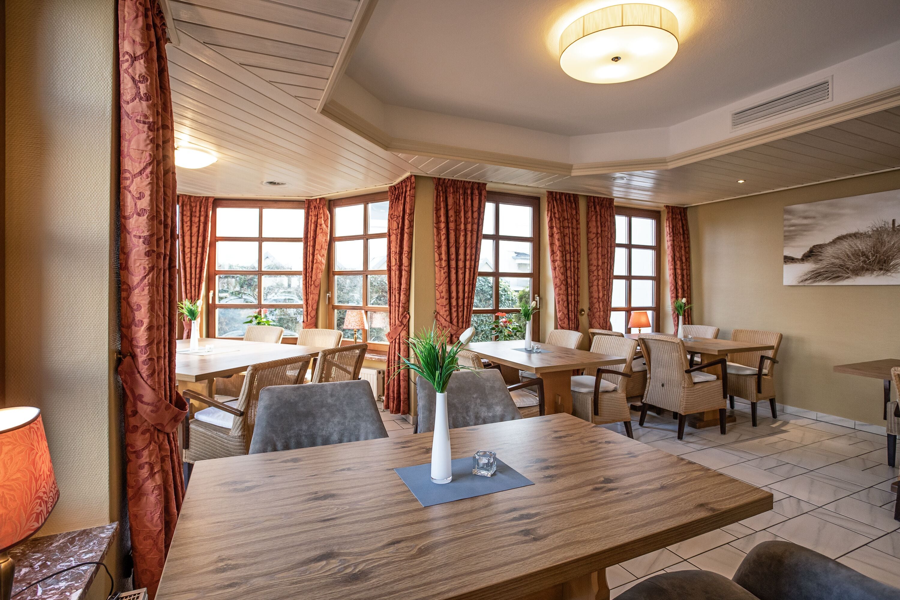 Comedor con mesas y sillas de madera, un florero con planta y amplias ventanas con cortinas.