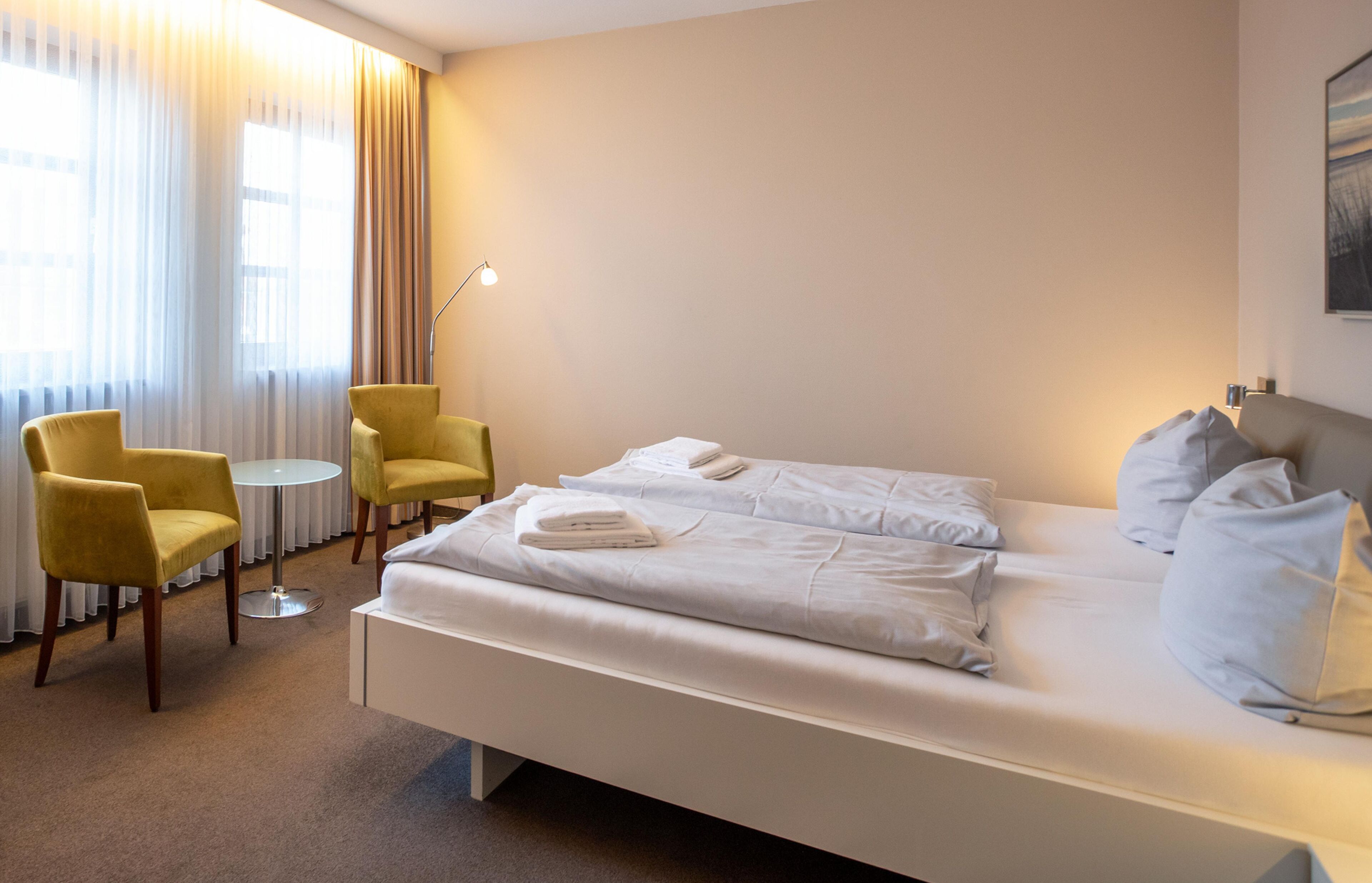 A hotel room with a bed, two yellow armchairs, a small round table, and a floor lamp.