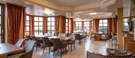 Un vaste espace dédié à la salle à manger, avec plusieurs tables et chaises, de grandes fenêtres dotées de rideaux et un ventilateur de plafond.
