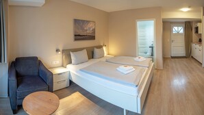 Ein Hotelzimmer mit einem Bett, einem Sessel, einem kleinen Tisch, einem Bild an der Wand und einer sichtbaren Küchenzeile durch eine offene Tür.