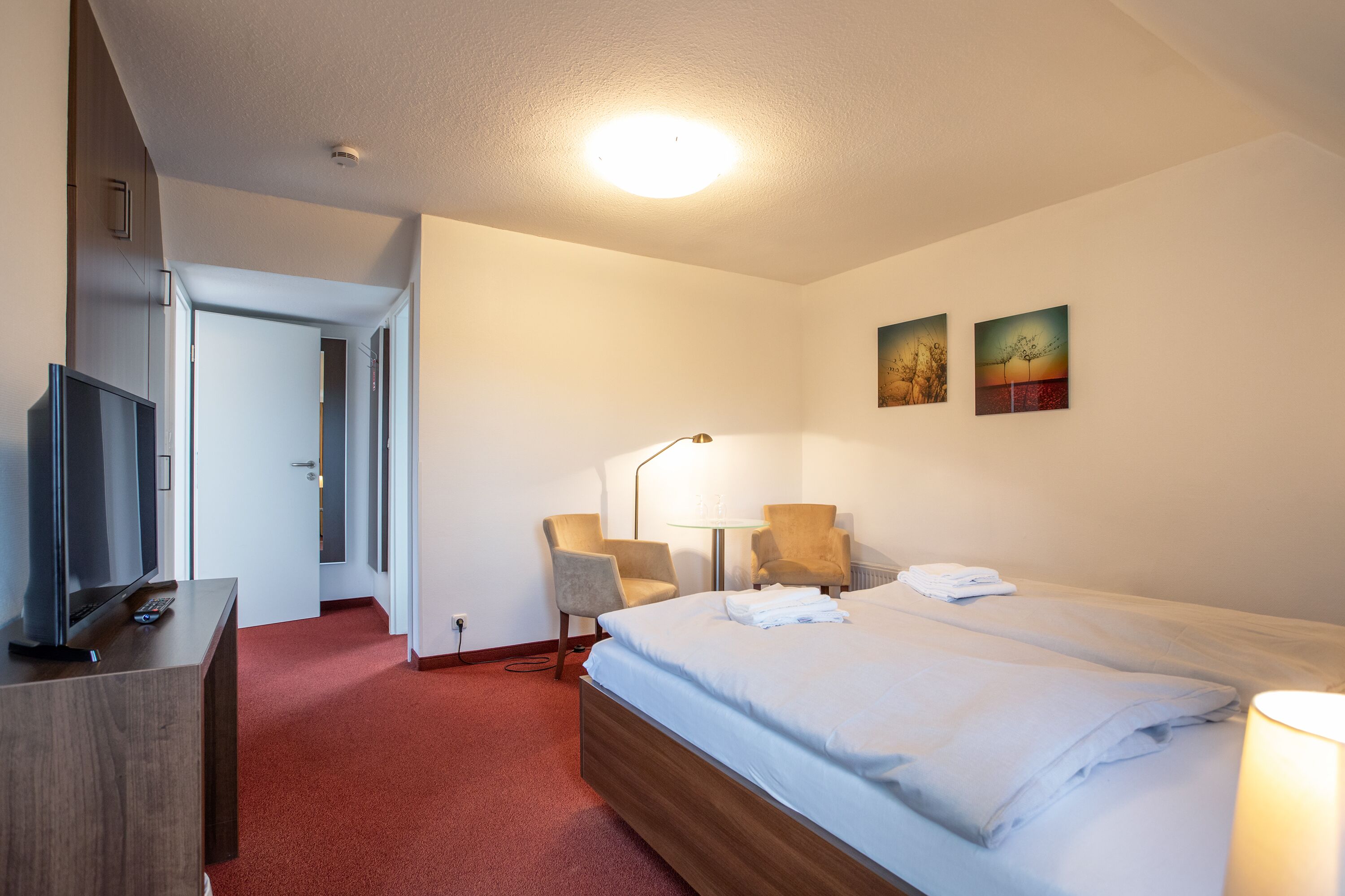 Una habitación de hotel con una cama, dos sillas, un televisor y obras de arte en la pared.