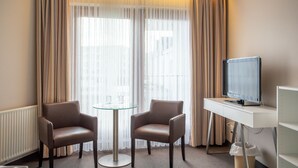 Ein Hotelzimmer mit Fernseher, zwei Ledersesseln, einem Glastisch und durchsichtigen Vorhängen.