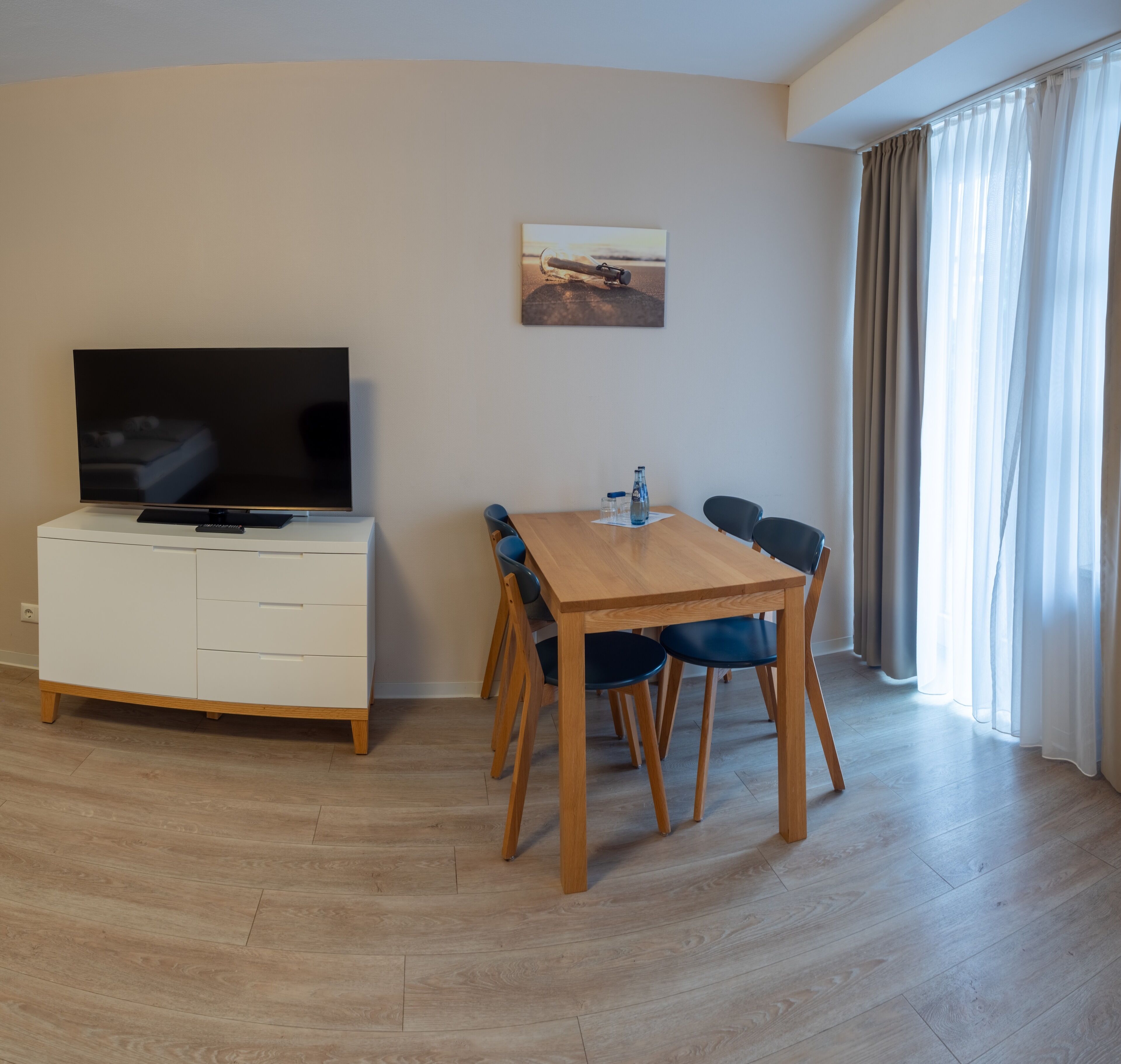 A modern living room with a flat-screen TV on a white cabinet, a wooden dining table with four chairs, and a large window with sheer curtains.