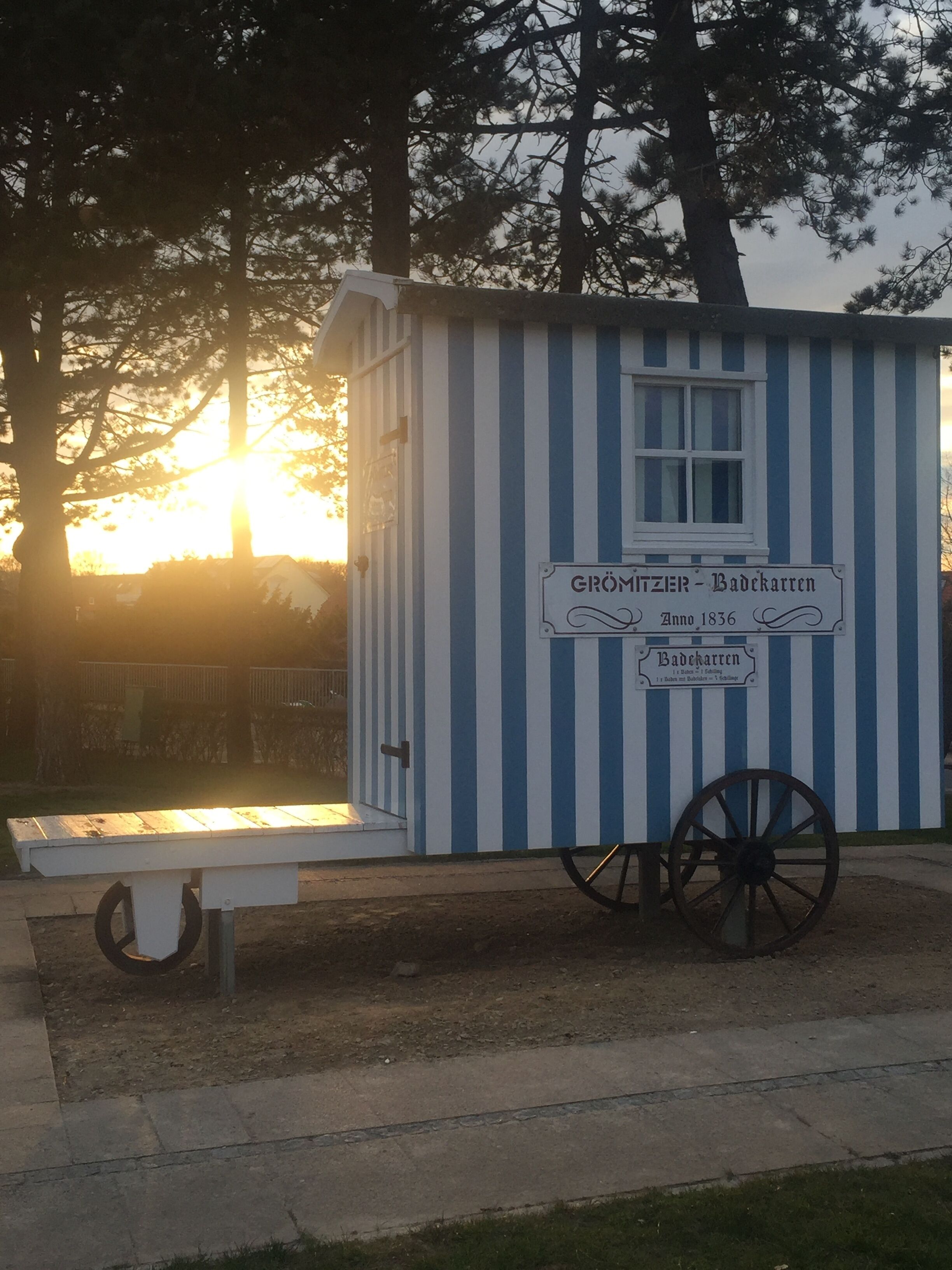 A small, single-story building with a blue and white striped exterior, a sign reading "Grömitzer Badekarren", and a wooden wheel attached to the front.