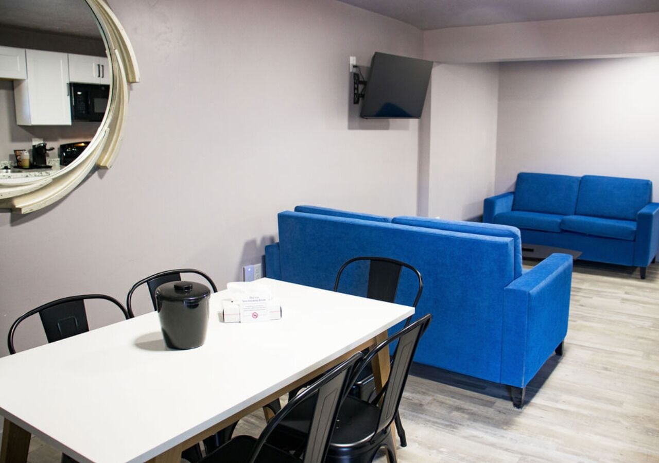 Una habitación con una mesa blanca, sillas negras, un sofá azul y un televisor montado en la pared.