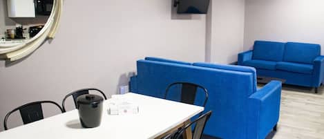 Una habitación con una mesa blanca, sillas negras, un sofá azul y un televisor montado en la pared.