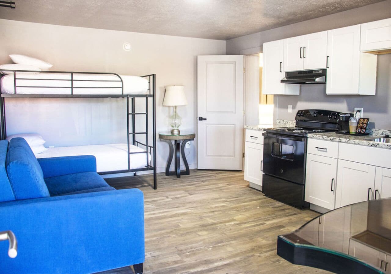 A compact room with a bunk bed, a blue sofa, a small table with a lamp, a kitchenette with a stove and oven, and white cabinets.