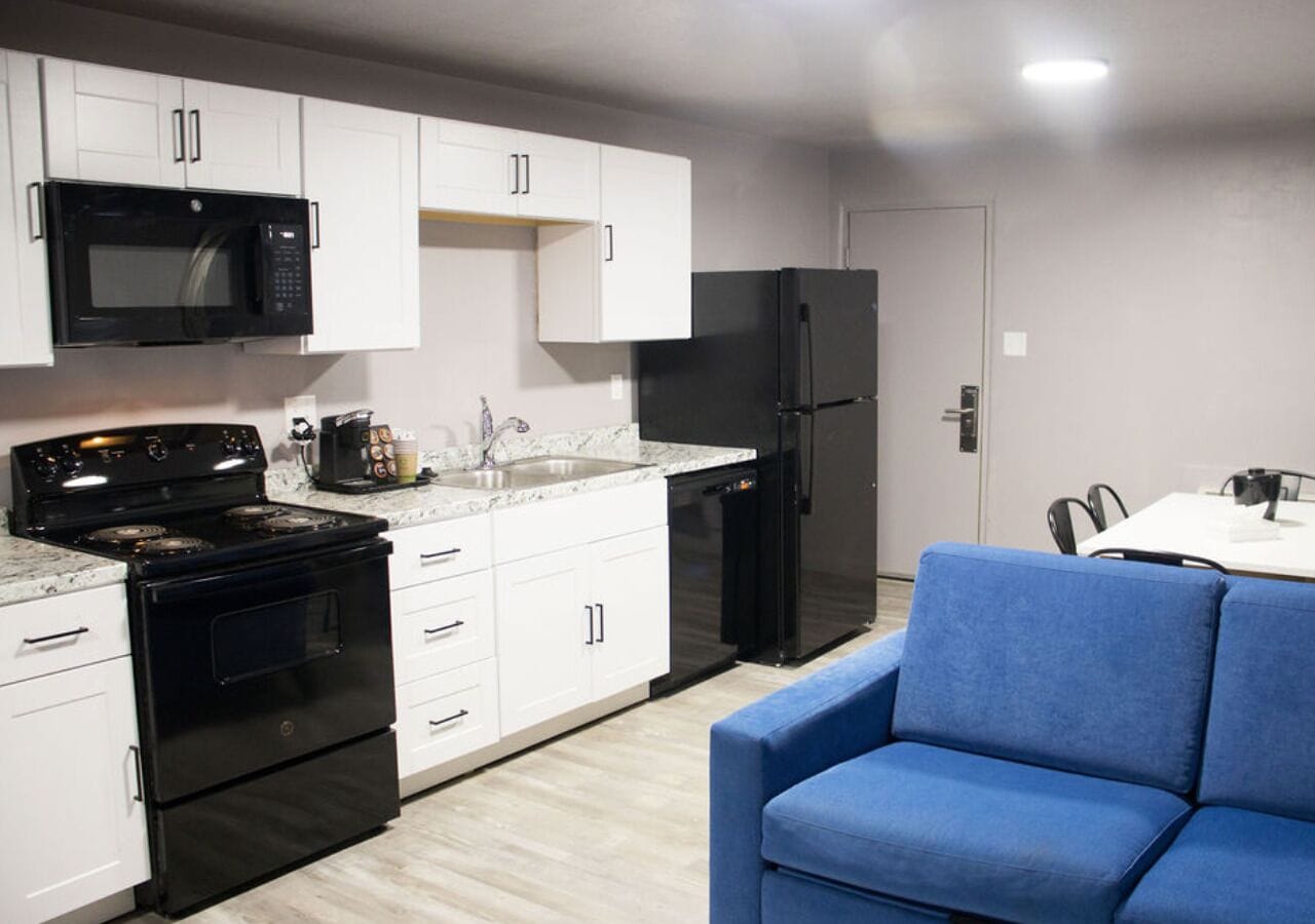 Cocina compacta con armarios blancos, un microondas negro, una estufa negra y un refrigerador negro. Hay un sofá azul y una pequeña mesa de comedor con sillas.