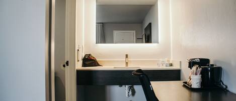 A modern bathroom with a large mirror, a sink, and a countertop with a coffee maker and a trash can.