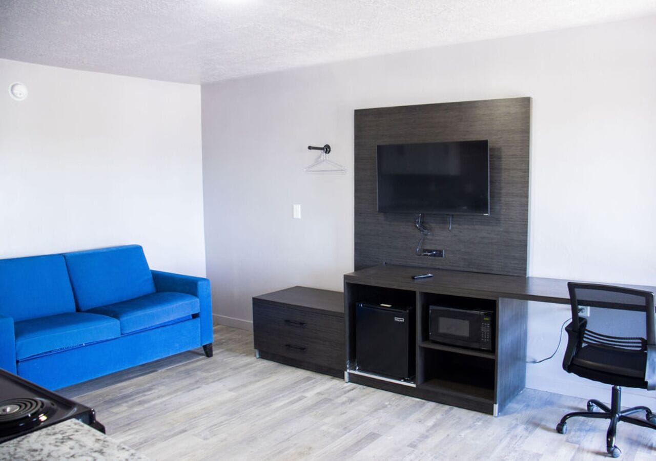 A modern living room with a blue sofa, a flat-screen TV mounted on a dark wood panel, a small black cabinet, and a black office chair.
