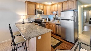A modern kitchen with wooden cabinets, stainless steel appliances, a granite countertop, and a bar stool. - Cute Ocean View Condo - Top Floor - Fireplace! (Lincoln City)