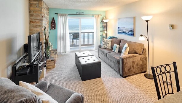 A living room with a TV, sofa, ottoman, and a balcony with a view. - Cute Ocean View Condo - Top Floor - Fireplace! (Lincoln City)
