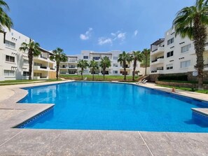 Una piscina rodeada de edificios modernos con balcones, palmeras y un cielo azul despejado.