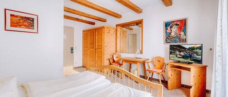 A bedroom with wooden beams, a bed, a desk with a chair, a television, and artwork on the walls.