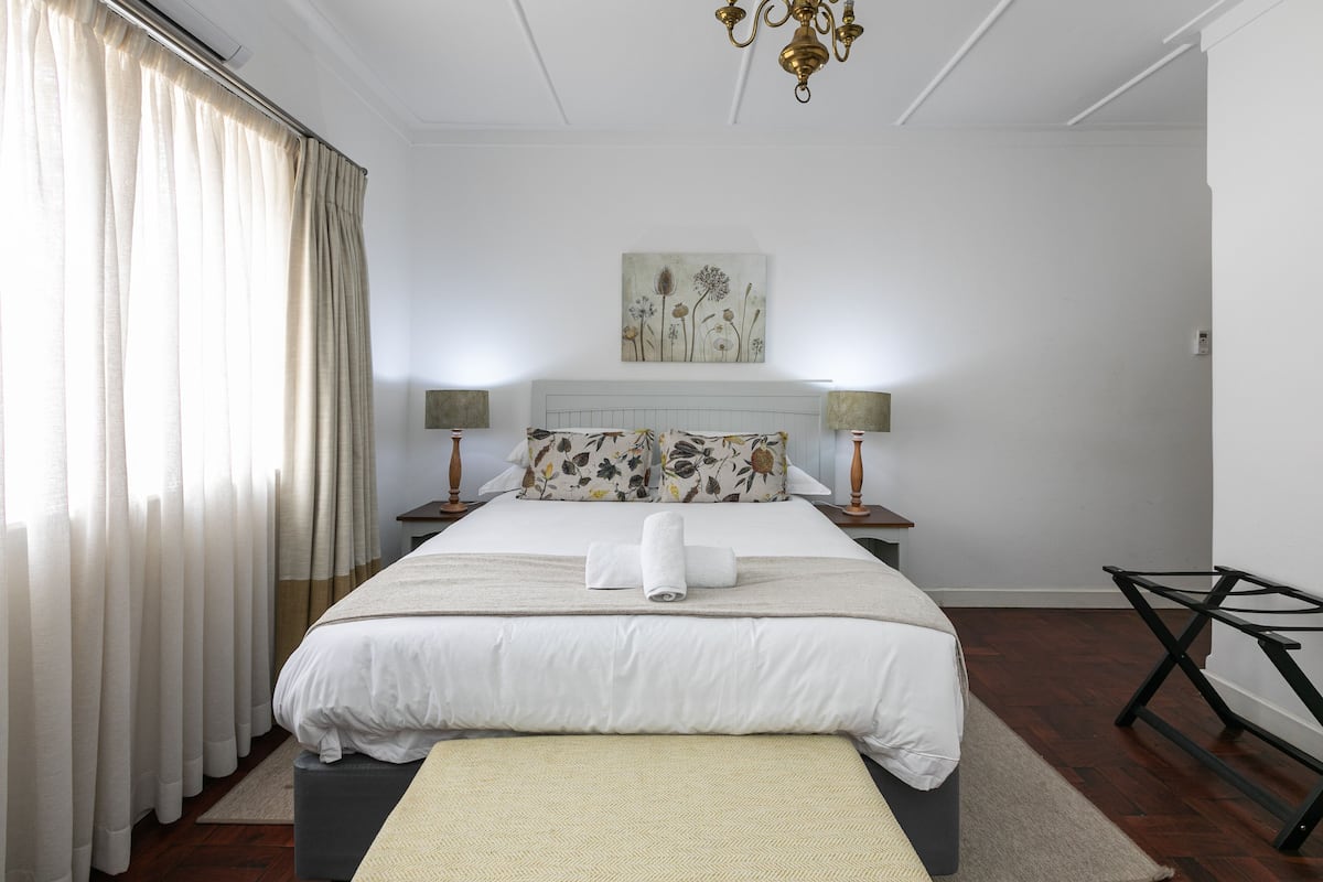 A neatly arranged bedroom with a large bed, a bedside table with a lamp, a wall-mounted artwork, and a modern chair.