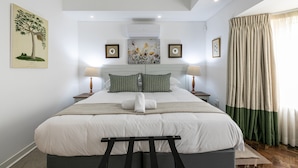A neatly arranged bedroom with a large bed, two bedside tables with lamps, a wall-mounted air conditioner, and a window with curtains. - Emmarentia Guest house  (Randburg)