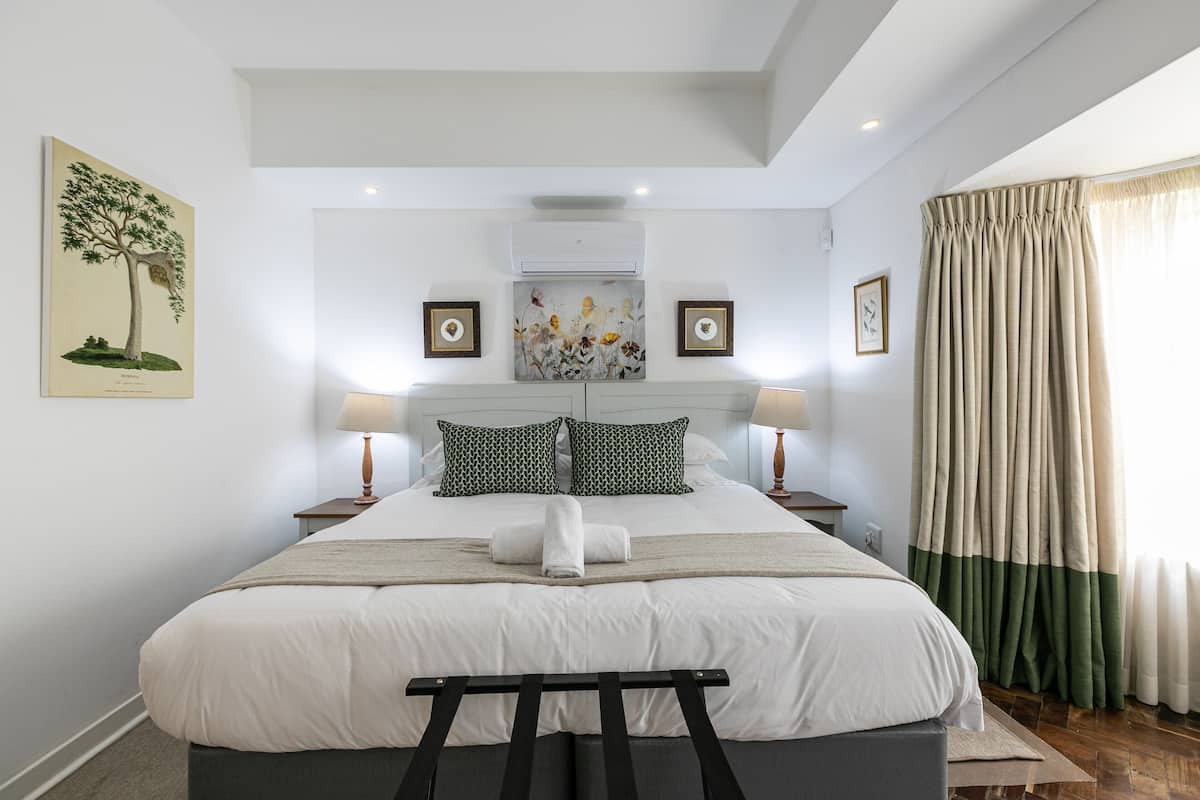 A neatly arranged bedroom with a large bed, two bedside tables with lamps, a wall-mounted air conditioner, and a window with curtains.
