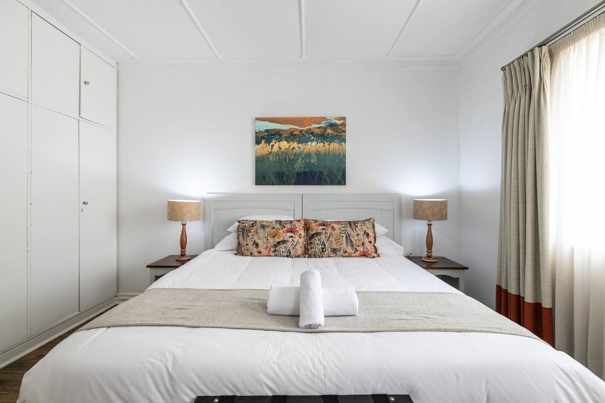 A neatly made bed with white linens, a decorative pillow, and a rolled towel on a beige rug.