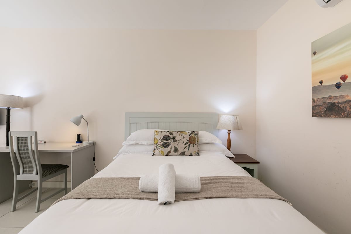 A neatly made bed with a rolled towel, a desk with a chair, and a lamp.