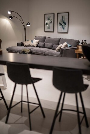 A modern living room with a grey sofa, black chairs, and a dining table. There are two wall-mounted lamps and abstract art pieces above the sofa.