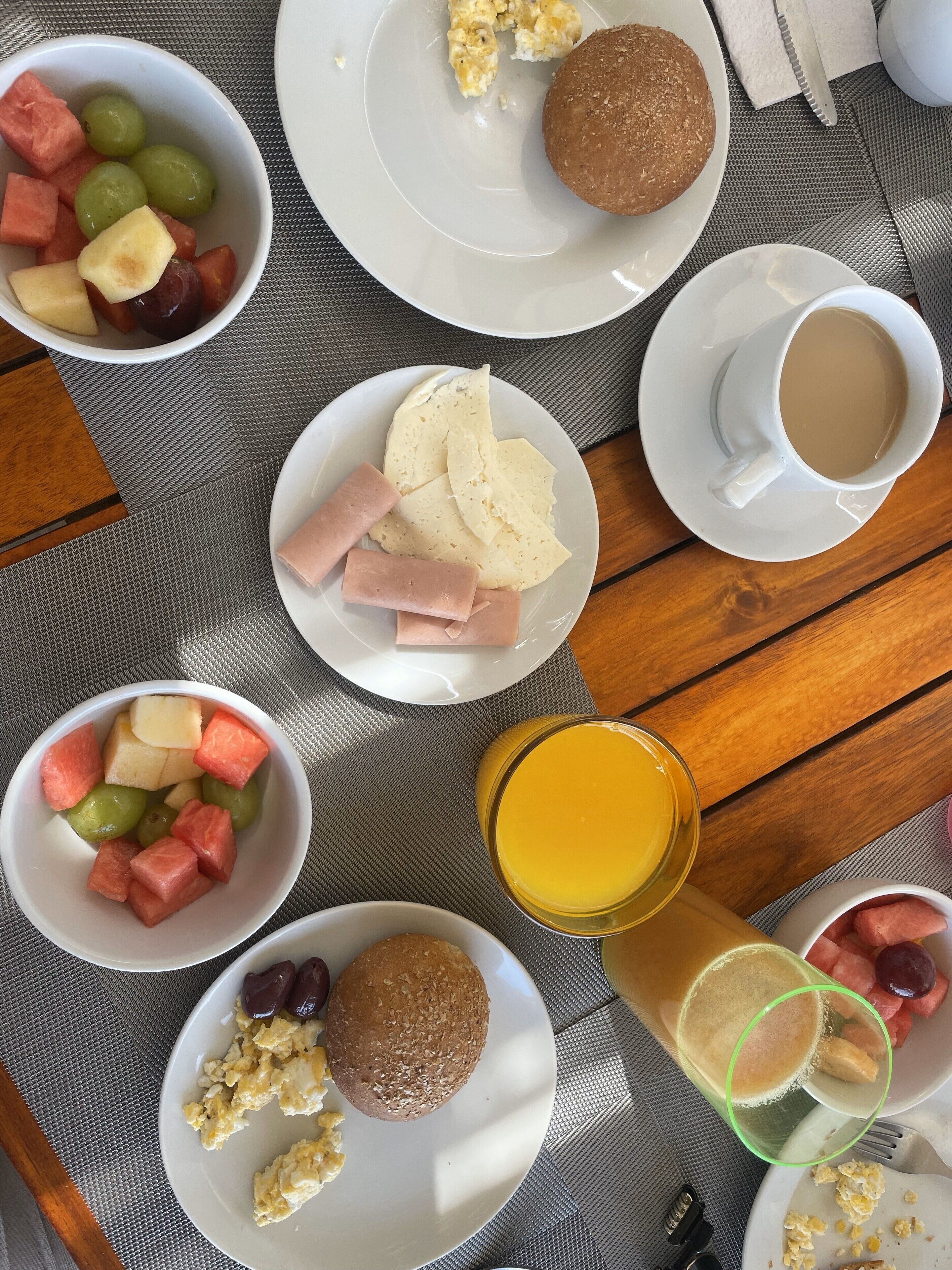 Mesa de café da manhã com ovos mexidos, torradas, frutas, uma xícara de café e um copo de suco de laranja.