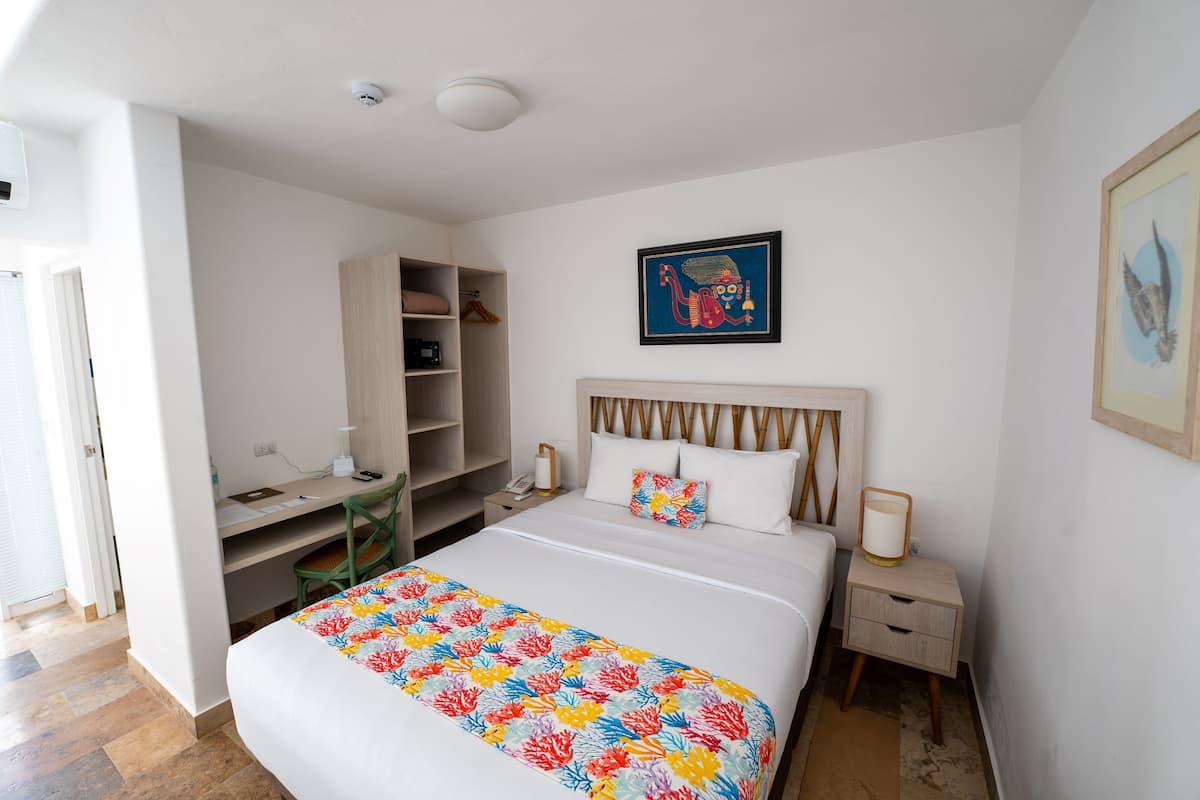 A hotel room with a bed, a desk, a chair, a wardrobe, and a nightstand with a lamp.