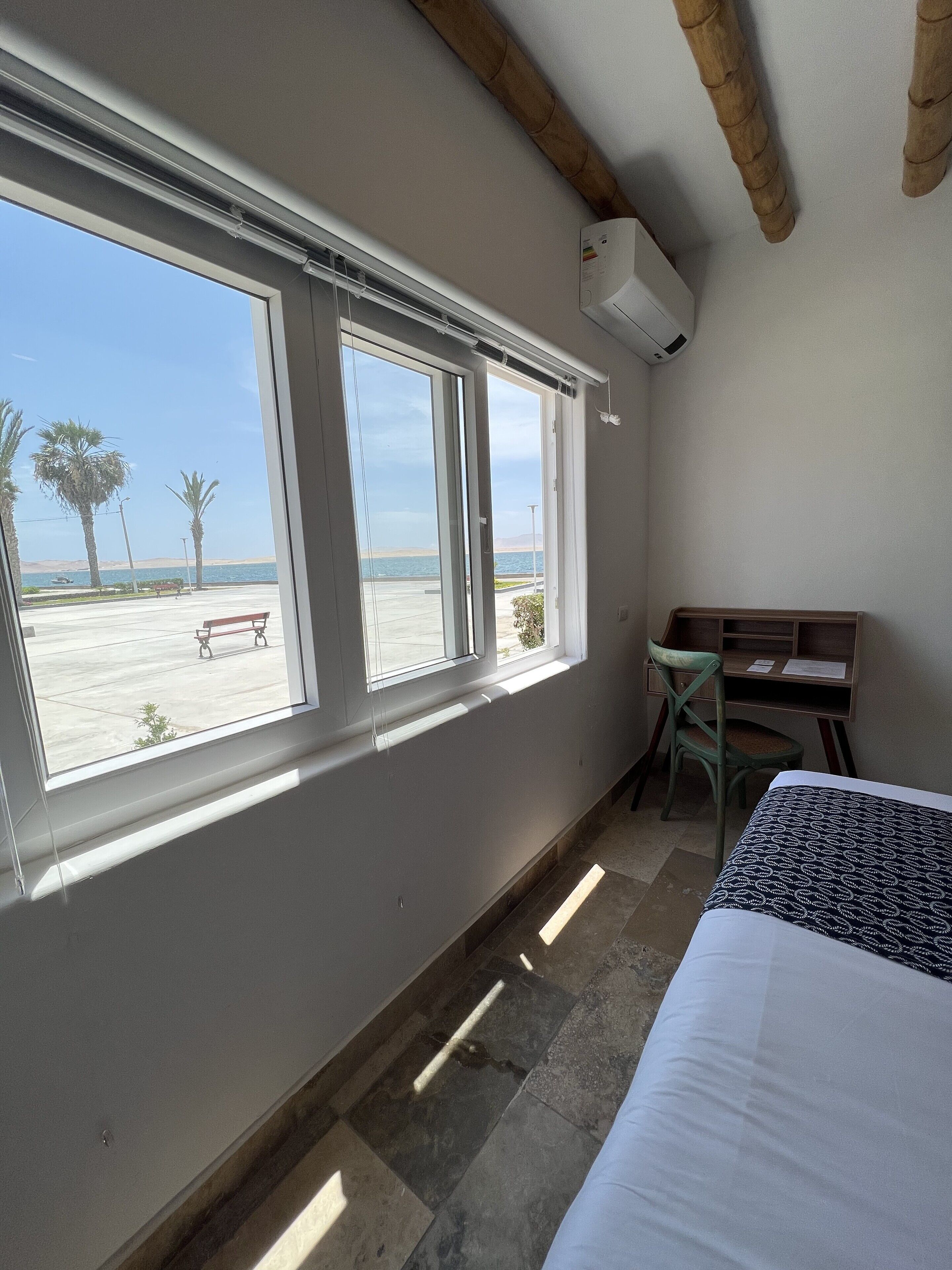 A room with a bed, a desk, and a window with a view of the beach and palm trees.