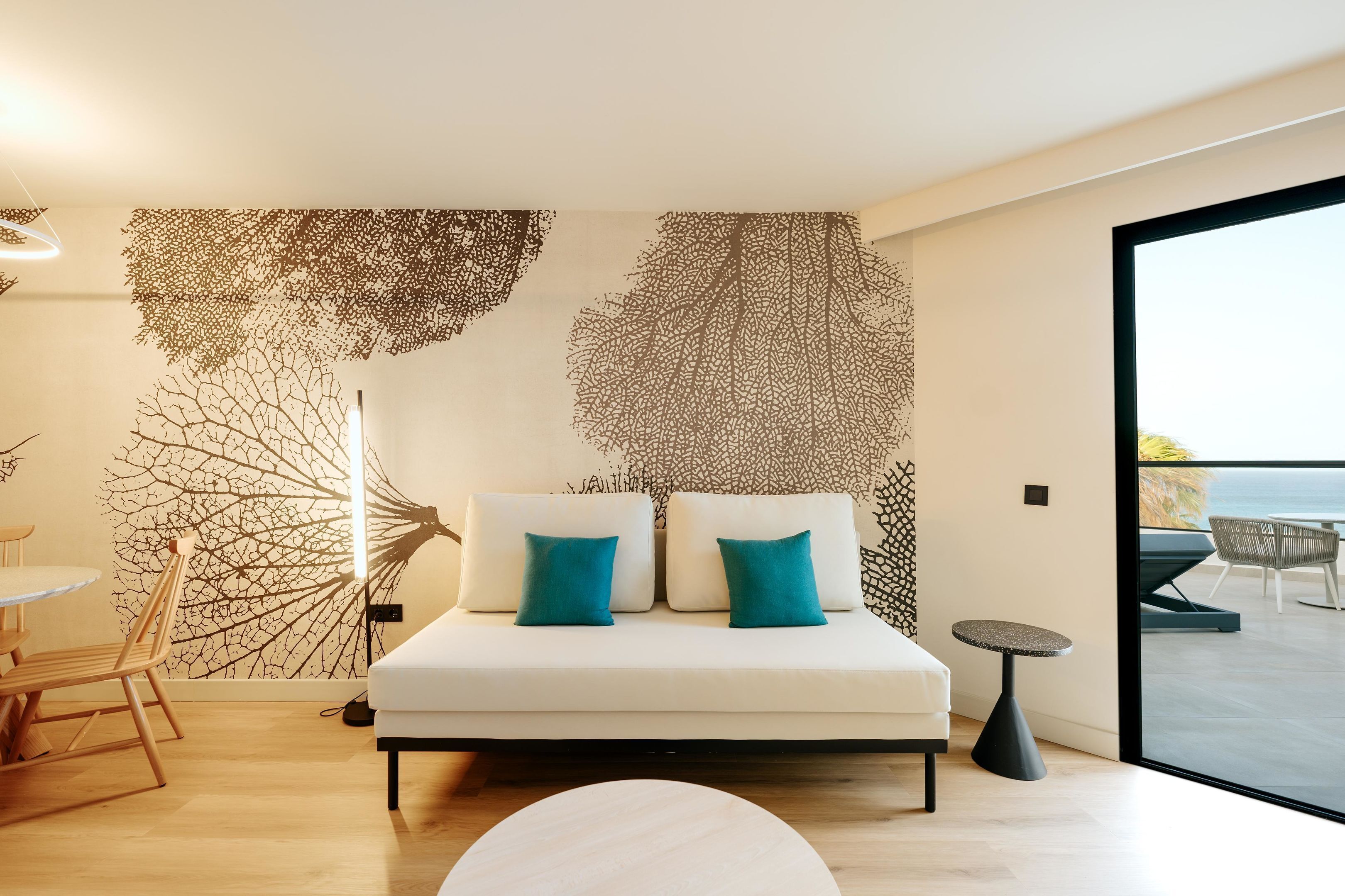 A modern bedroom with a large bed, two teal pillows, a round side table, and a view of the sea through a large window.