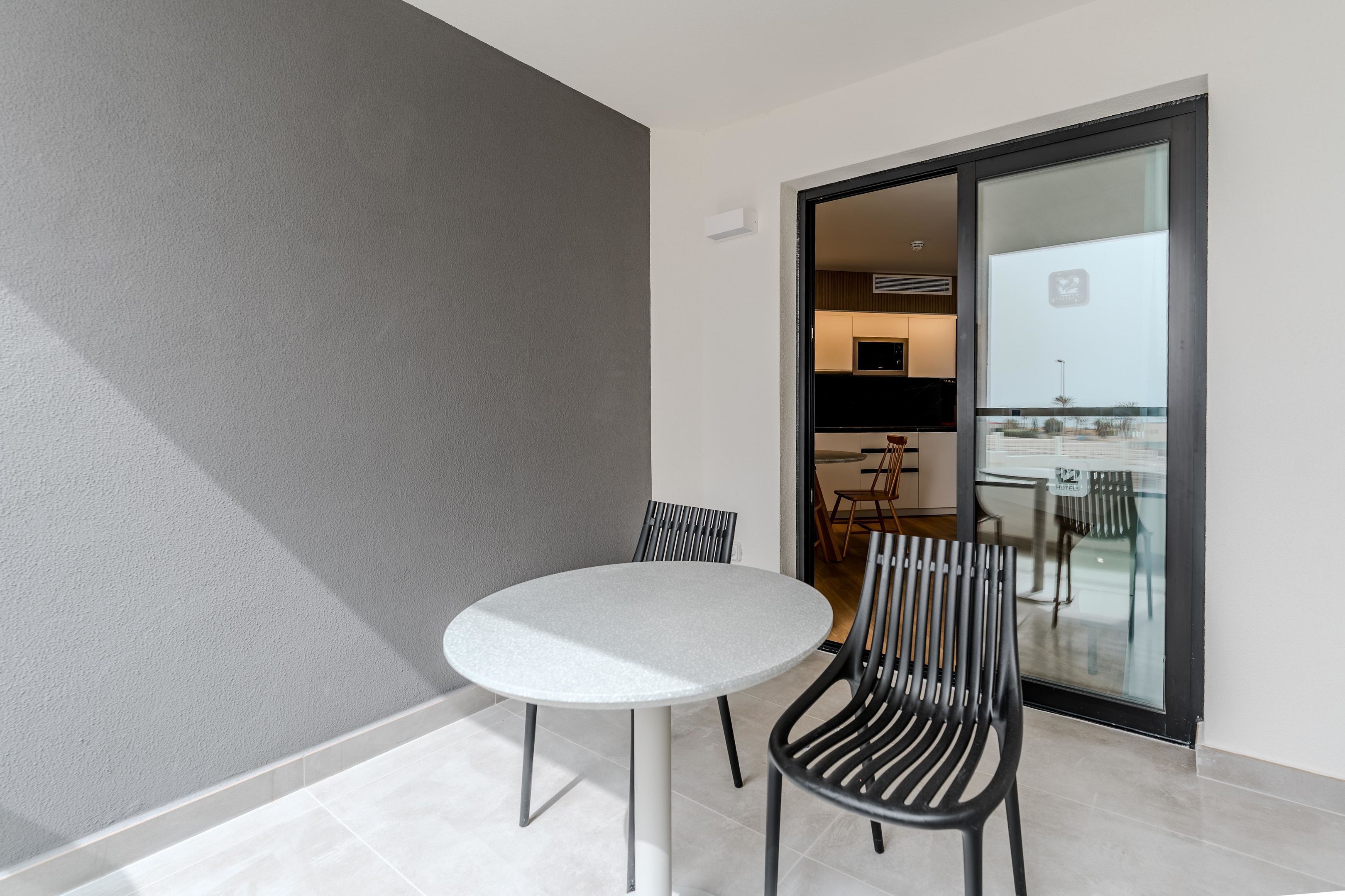A modern dining area with a round table and black chairs, a grey wall, and a sliding glass door leading to a kitchen.