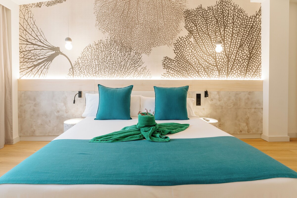 A hotel room with a large bed, two pillows, a green throw blanket, and a wall with a leaf pattern.