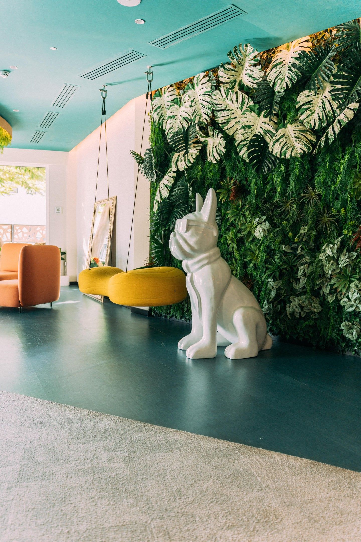 Een modern interieur met een witte konijnenbeeld, een groene wand met tropische begroeiing en oranje en gele zitplaatsen.