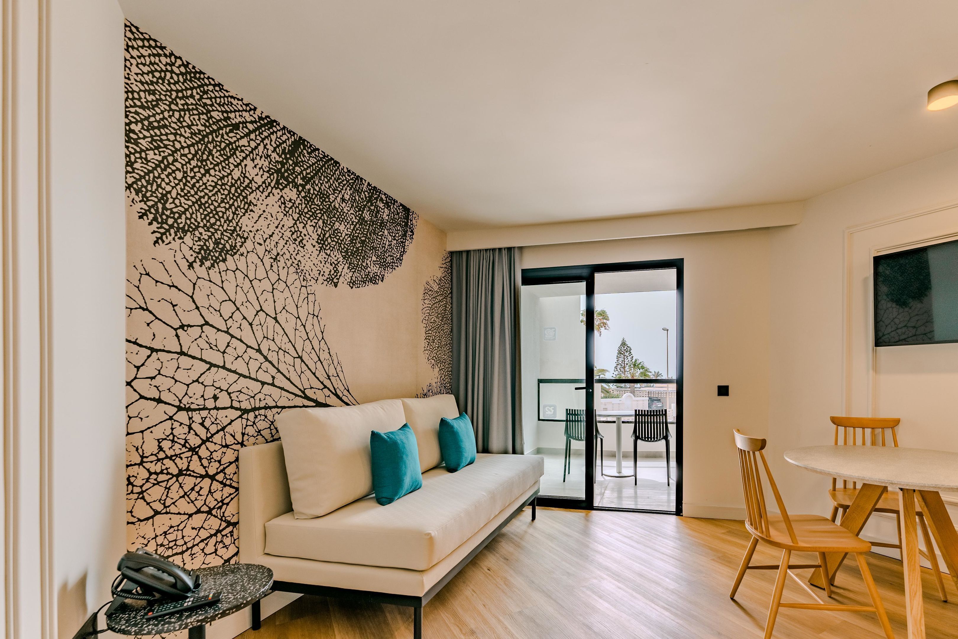 Ein modernes Wohnzimmer mit einem weißen Sofa, türkisfarbenen Kissen, einer Wand mit Muster und einem Esstisch mit Holzstühlen, sowie einer Glasschiebetür zu einem Außenbereich.