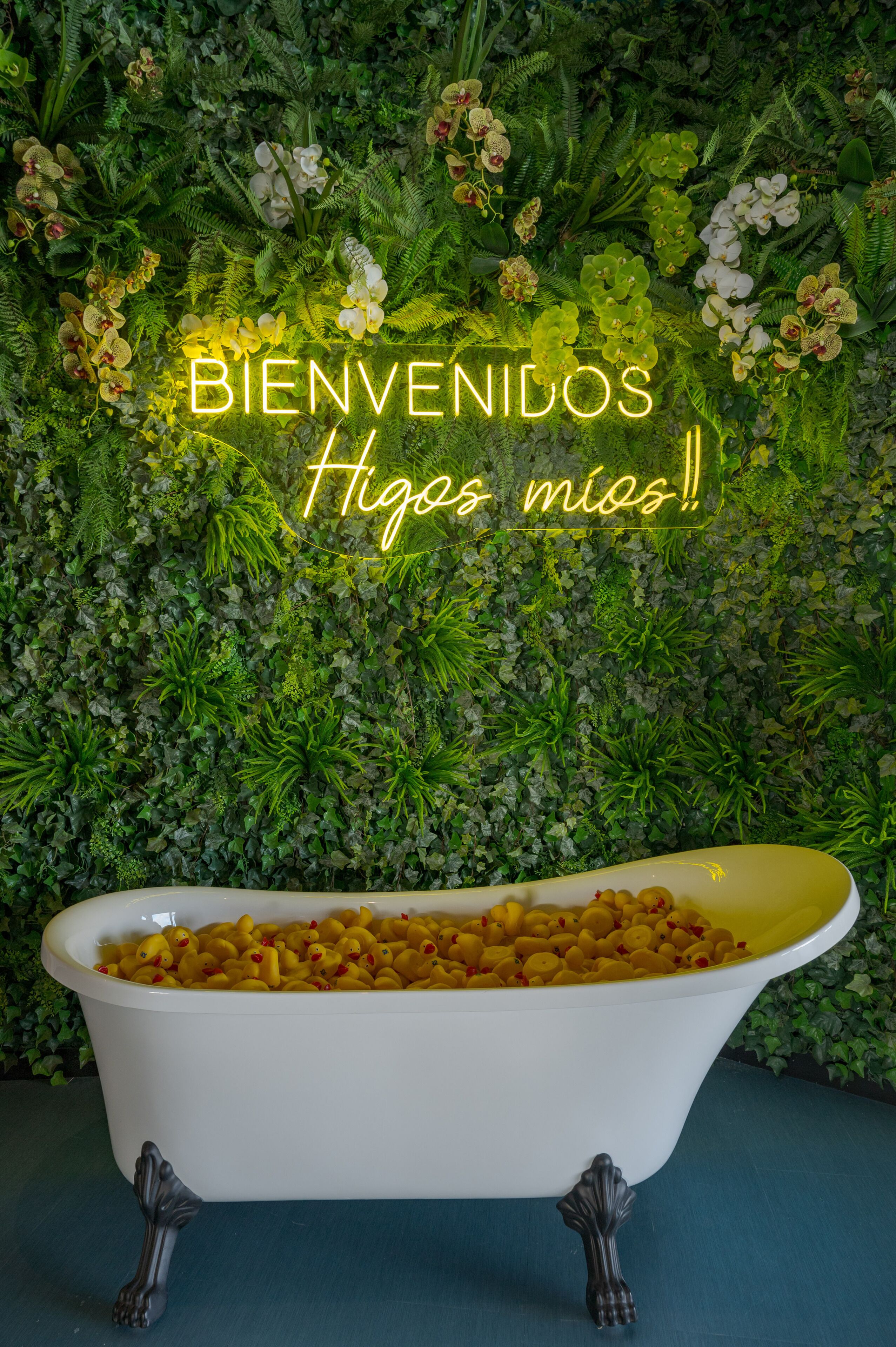Una bañera blanca llena de juguetes de baño amarillos, frente a una pared verde exuberante con diversas plantas y flores.
