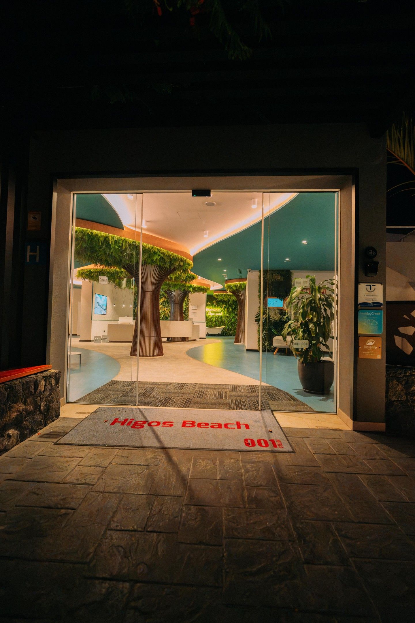 A hotel entrance with a reception desk, a green wall with plants, and a sign reading 'Higos Beach 001'.