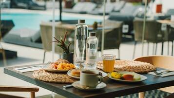 A table with breakfast items, a bottle, and a glass of juice in a poolside setting.