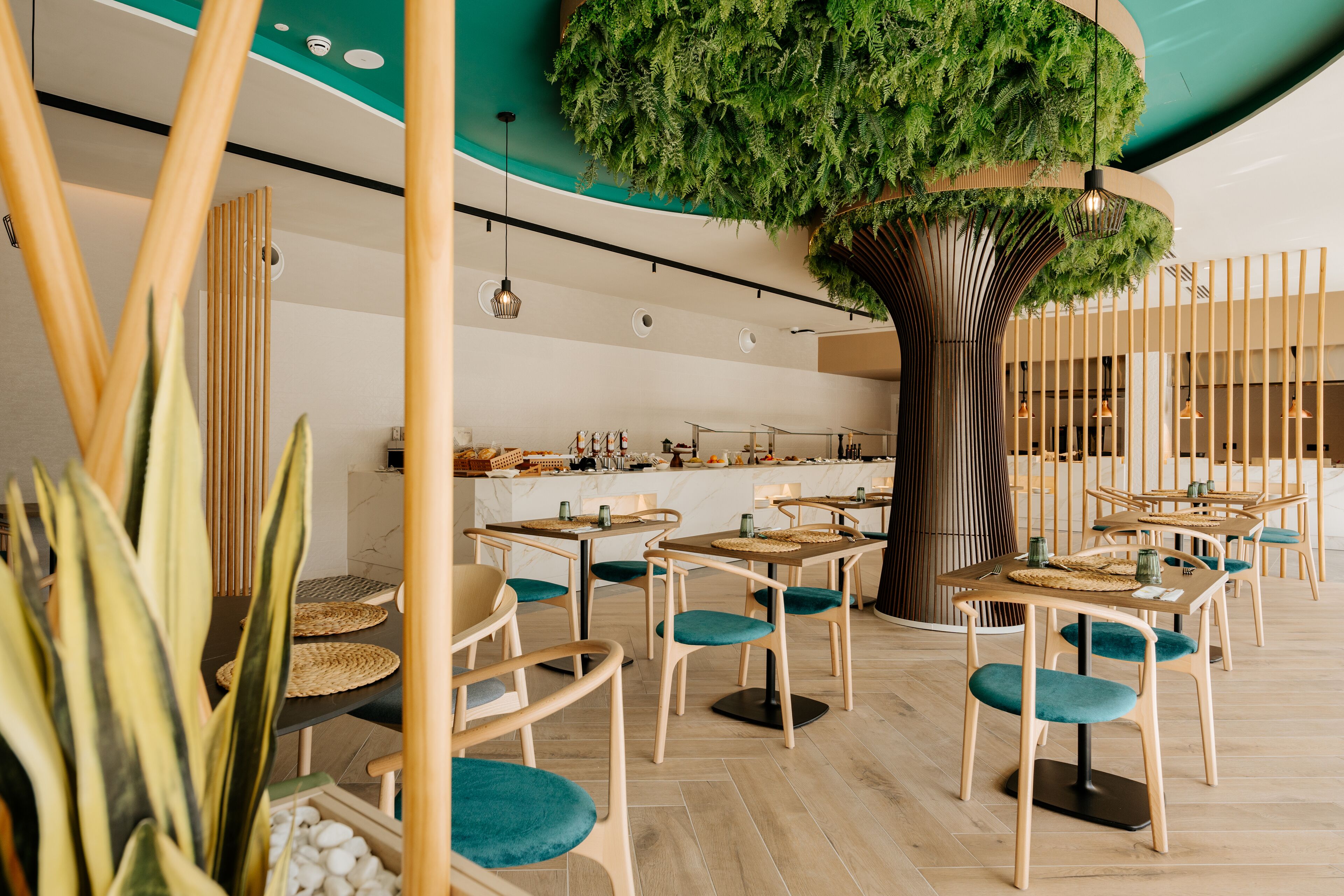 A modern restaurant with wooden tables and teal chairs, a central tree, and a ceiling with a green leaf design.