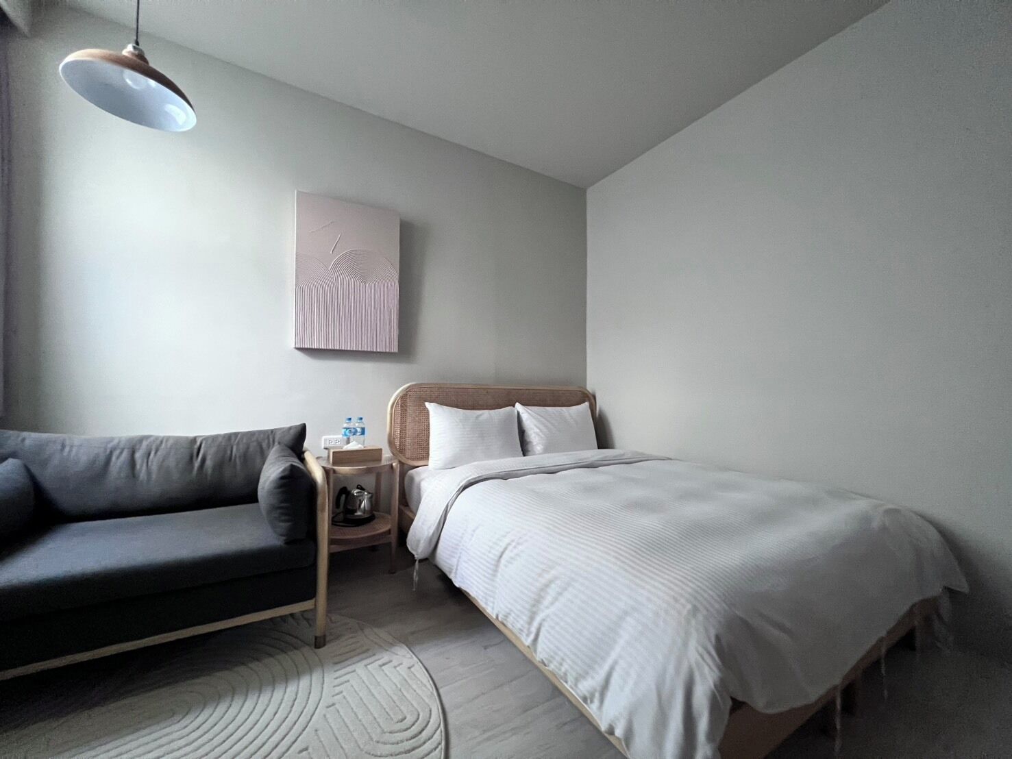 A minimalist bedroom with a bed, a sofa, a bedside table with a lamp, and a wall art piece.