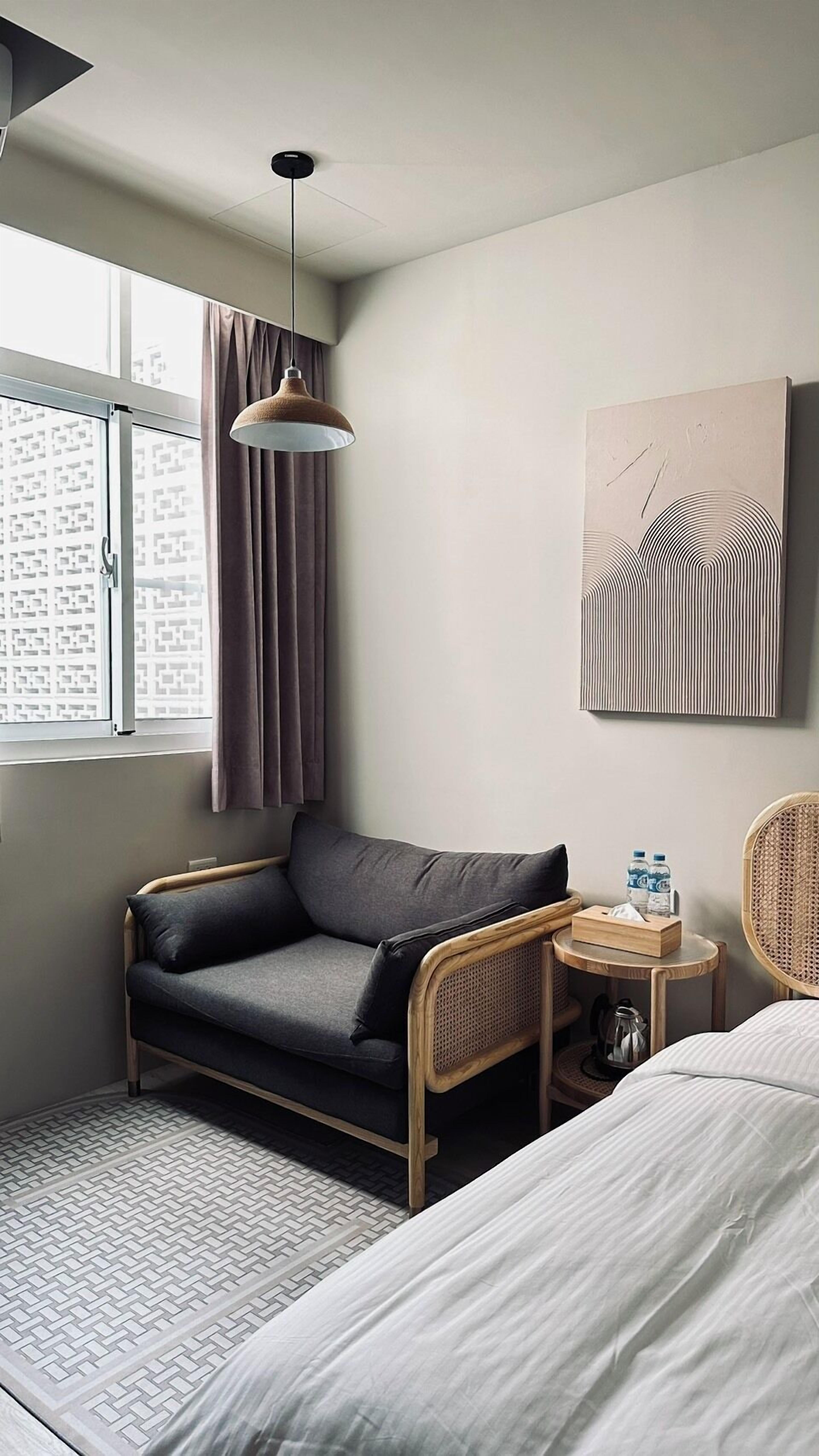 A modern hotel room with a sofa, a small table with a bottle and tissues, a wicker chair, a bed, and a large window with curtains.