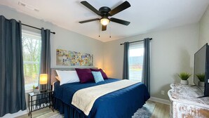 Een slaapkamer met een bed, een plafondventilator, een raam met gordijnen, een nachtkastje met een lamp en een dressoir.