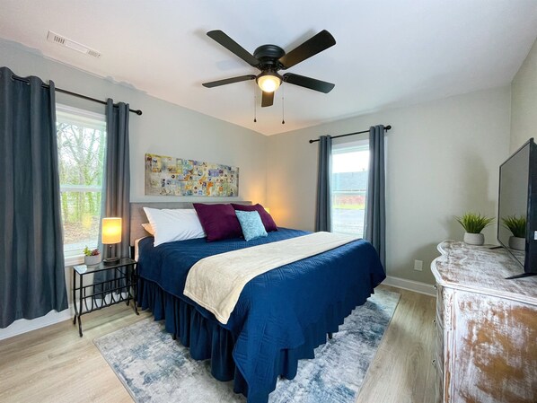 A bedroom with a bed, a ceiling fan, a window with curtains, a bedside table with a lamp, and a dresser.