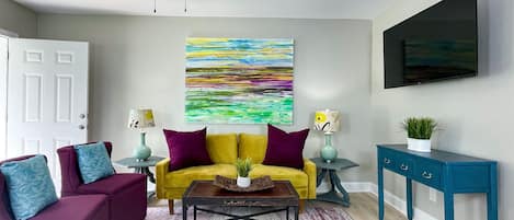A living room with a yellow sofa, purple armchairs, a coffee table, a ceiling fan, a painting, a TV, and a blue console table.