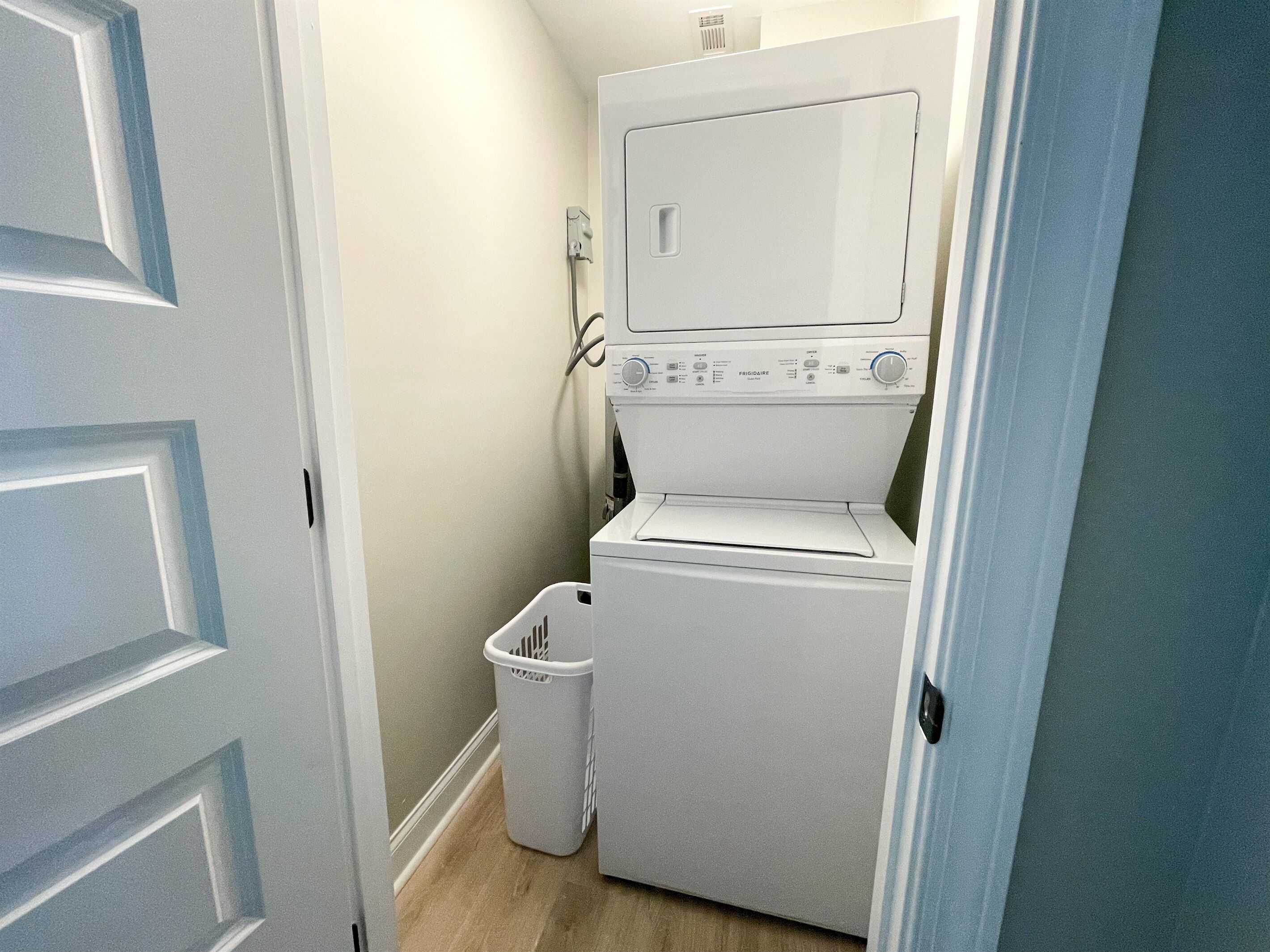 Een kleine wasruimte met een voorlader wasmachine en droger, een kleine vuilnisbak en een deur naar een andere ruimte.