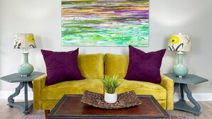Een kamer met een gele bank, paarse kussens, twee nachtkastjes met lampen, een salontafel met een plant en een kleurrijk abstract schilderij aan de muur.