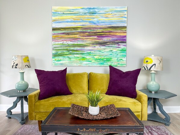 Una habitación con un sofá amarillo, cojines morados, dos mesitas de noche con lámparas, una mesa de centro con una planta y un cuadro abstracto y colorido en la pared.