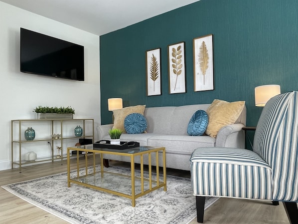 Ein modernes Wohnzimmer mit einer Türkis-farbenen Akzentwand, einem grauen Sofa, einem gestreiften Sessel und einem Couchtisch mit goldener Rahmen.