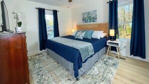 Un dormitorio con cama, ventilador de techo, mesita de noche, lámpara y ventana con cortinas.