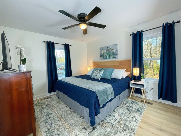 A bedroom with a bed, a ceiling fan, a nightstand, a lamp, and a window with curtains. - NEW w/ King Bed Near Tennis Center & Berry College (Rome)