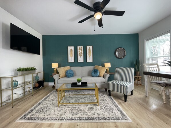 A living room with teal walls, a ceiling fan, a flat-screen TV, a sofa with decorative pillows, a striped armchair, a coffee table, and a patterned rug. - NEW w/ King Bed Near Tennis Center & Berry College (Rome)